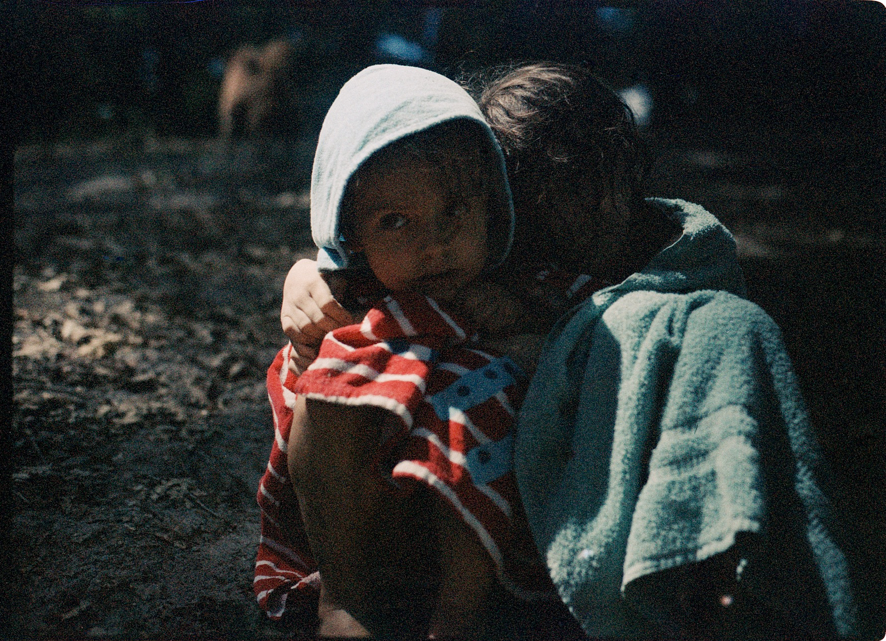 Eine Analogfotografie mit ausgeprägtem Filmkorn, auf dem zwei Kinder im Halbschatten sitzend zu sehen sind, eingehüllt in Handtücher und eng umarmend.